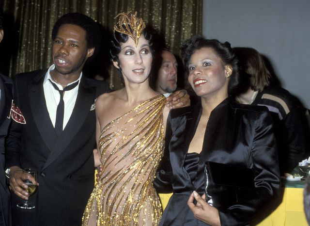 A photo of Nile Rodgers in a black suit, Cher in a golden nude gown and headpiece, and Alfa Anderson in a black dress and jacket